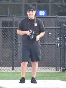 Junior Alex Duley stands on Heartland Farms Field at Amherst High School, holding a camera in his hands.
